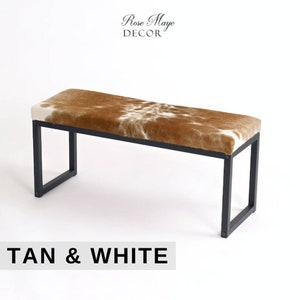 May include: Tan and white cowhide bench with a black metal frame. The bench is a simple design with a rectangular seat and four legs.