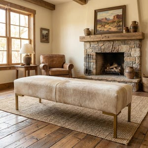 May include: A rectangular bench upholstered in light beige and cream-colored cowhide leather, featuring a smooth, hair-on-hide texture. The bench is supported by four straight, metallic gold-toned legs with a clean, angular design.
