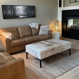 May include: A living room with a brown leather sofa, a white and brown cowhide ottoman, and a jute rug. The sofa has two throw pillows and a throw blanket. The ottoman is in front of the sofa and is centered on the rug. The rug is on a hardwood floor. There is a black fireplace with a black tile surround in the background.