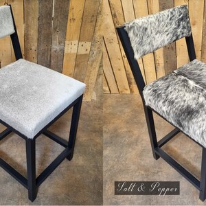 May include: Two black metal bar stools with a square base. The stool on the left has a gray cowhide seat and back. The stool on the right has a black and white cowhide seat and back. The stools are labeled "Grey" and "Salt & Pepper".