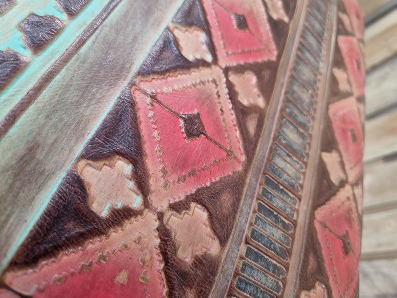 May include: Close-up of a brown leather bag with a geometric pattern in pink, white, and turquoise. The pattern is stitched into the leather and has a rustic, vintage look.