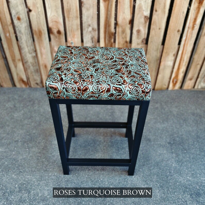 Op de afbeelding: Een zwarte metalen barstoel met een turquoise en bruine bloemenpatroon op de zitting. De zitting heeft een reli&euml;fpatroon van rozen. De stoel heeft een vierkante basis met een enkele dwarsbalk.