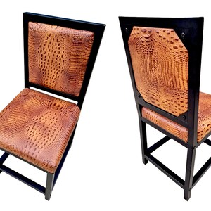 May include: Two black metal dining chairs with brown crocodile-embossed leather upholstery. The chairs have a square back and a simple, sturdy design.