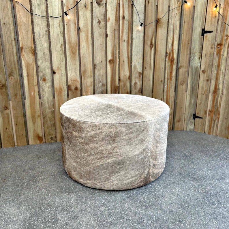 May include: A round, light brown, cowhide ottoman with a textured surface. The ottoman is sitting on a gray concrete floor.