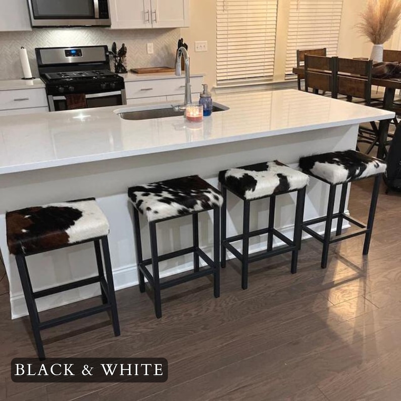 May include: Four black metal bar stools with cowhide seat cushions in black and white. The stools are arranged in a row in front of a white kitchen island.