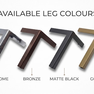 May include: Four L-shaped furniture legs arranged in a row, labeled with the text AVAILABLE LEG COLOURS. The finishes from left to right are shiny chrome, dark bronze, matte black, and gold. Each leg features a two-pronged design.
