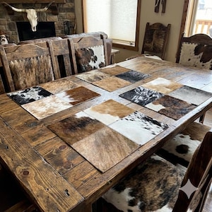 May include: A wooden dining table with matching chairs, featuring cowhide patchwork placemats. The placemats display a variety of colours and patterns. The chairs are upholstered in cowhide, creating a rustic, country-style dining set.