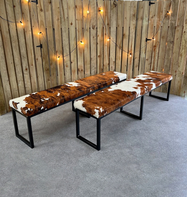 May include: Two black metal framed benches with brown and white cowhide upholstery. The benches are set against a wood slat wall with string lights.