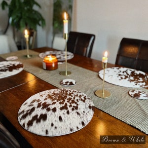 Puede incluir: Una mesa de comedor con manteles individuales y posavasos de piel de vaca marrón y blanca. Candelabros dorados con velas encendidas se colocan sobre un camino de mesa marrón. El texto "Brown & White" está en la parte inferior.