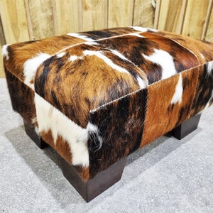 May include: A brown, white, and black cowhide ottoman with a square top and four wooden legs.