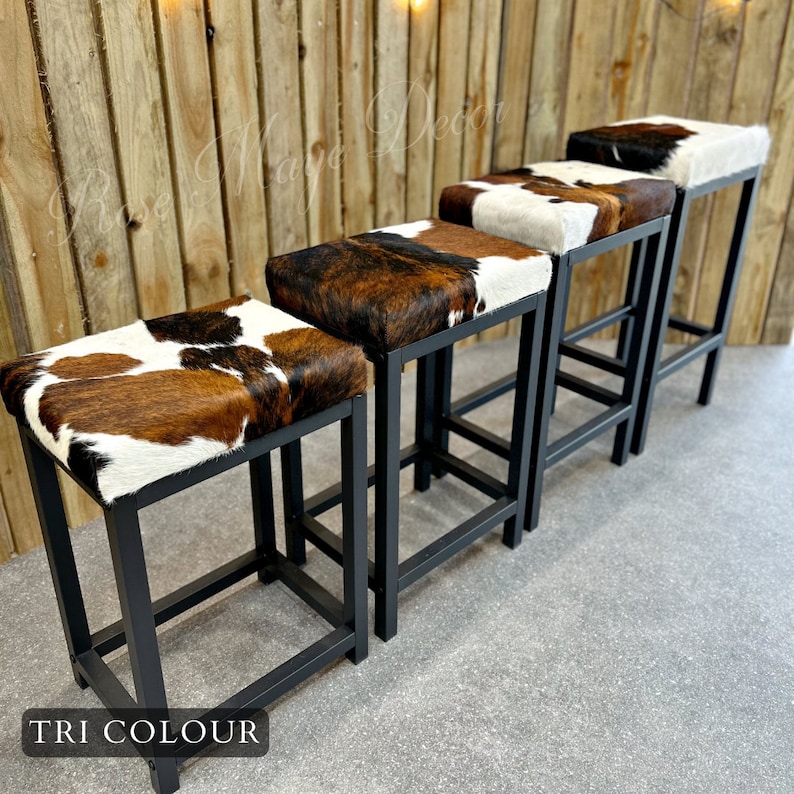 May include: Four black metal bar stools with brown, white, and black cowhide seats. The stools are arranged in a row, with the front stool closest to the viewer. The stools are in front of a wooden wall.