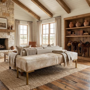 May include: A rectangular bench upholstered in light beige cowhide with a smooth texture, supported by four metallic gold legs. The bench is positioned in front of a sofa draped with a neutral-toned throw blanket in a room with wooden beams.