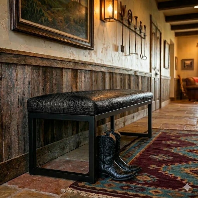 Op de afbeelding: Een zwarte bank met een imitatie-alligatorhuid bovenkant en een zwart metalen frame. Twee zwarte cowboylaarzen staan onder de bank. De bank staat in een gang met een rustieke houten wand.