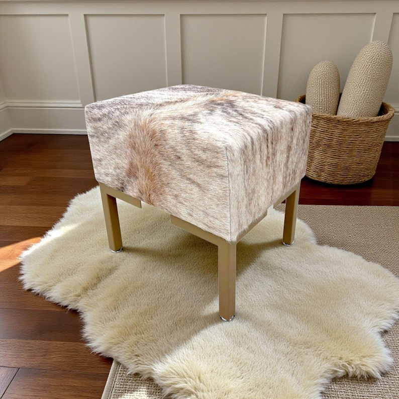 May include: A square ottoman with a cowhide pattern in shades of brown, tan, and white. The ottoman has a light gold metal frame and is placed on a cream-colored sheepskin rug. The background includes a woven basket and a light-colored wall.