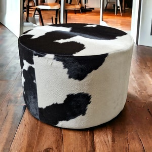May include: A round, black and white cowhide ottoman with a soft, plush texture. The ottoman is sitting on a wooden floor.