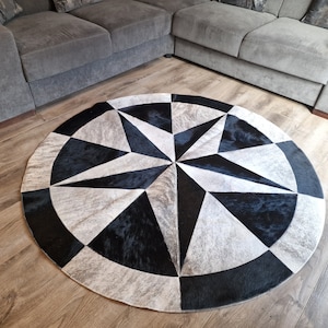 May include: Round cowhide rug with a geometric starburst design. The rug features black, white, and gray sections arranged in a radial pattern. The rug is placed on a wooden floor, near a gray sectional sofa.