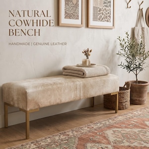 May include: A rectangular bench upholstered in light beige natural cowhide leather with four metallic gold legs. The text NATURAL COWHIDE BENCH HANDMADE | GENUINE LEATHER is visible in the upper left corner above the bench.