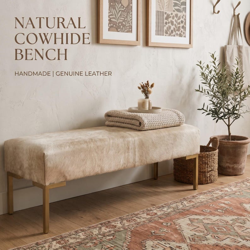May include: A rectangular bench upholstered in light beige natural cowhide leather with four metallic gold legs. The text NATURAL COWHIDE BENCH HANDMADE | GENUINE LEATHER is visible in the upper left corner above the bench.