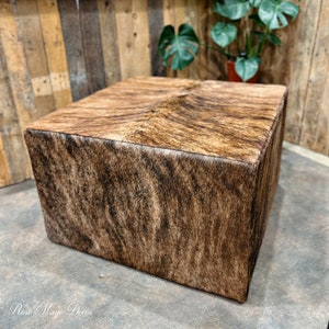 Pu&ograve; includere: Un pouf quadrato rivestito in pelle di mucca marrone e nera con un leggero bagliore. Il pouf &egrave; appoggiato su un pavimento in cemento grigio.
