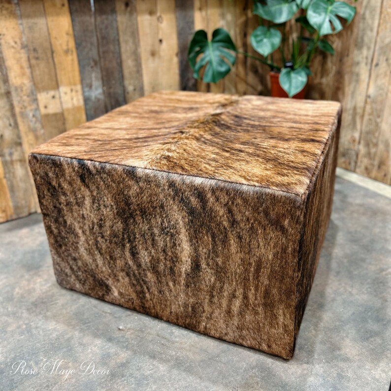 Pu&ograve; includere: Un pouf quadrato rivestito in pelle di mucca marrone e nera con un leggero bagliore. Il pouf &egrave; appoggiato su un pavimento in cemento grigio.