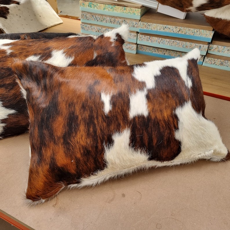 Puede incluir: Dos almohadones de piel de vaca marr&oacute;n, blanco y negro con una textura suave y peluda.