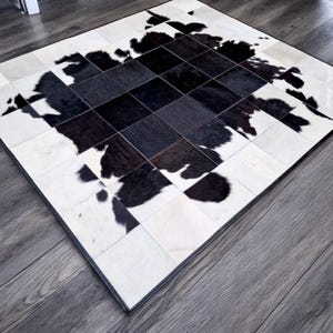Könnte beinhalten: Ein quadratischer Kuhfellteppich mit Patchwork-Design. Der Teppich weist ein Muster aus schwarzen und weißen Quadraten und unregelmäßigen Formen auf, das einen auffälligen Kontrast erzeugt. Der Teppich liegt auf einem Holzboden.
