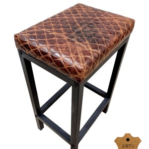 May include: A brown leather bar stool with a diamond stitch pattern. The stool has a black metal frame and a 100% genuine leather seat. The stool is perfect for adding a touch of rustic charm to any bar or kitchen.
