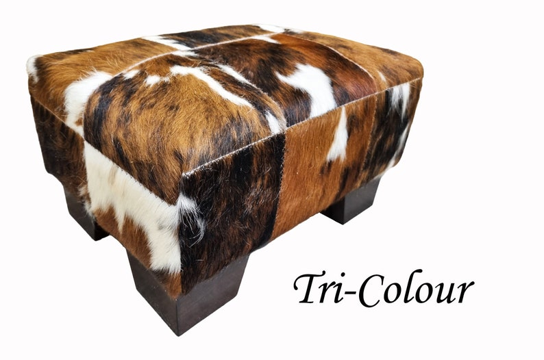 May include: A brown, white, and black cowhide ottoman with wooden legs. The ottoman is labeled "Tri-Colour".
