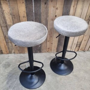 Op de afbeelding: Twee grijze en witte barstoelen met een zwart metalen onderstel en voetsteunen. De krukken zijn in hoogte verstelbaar.