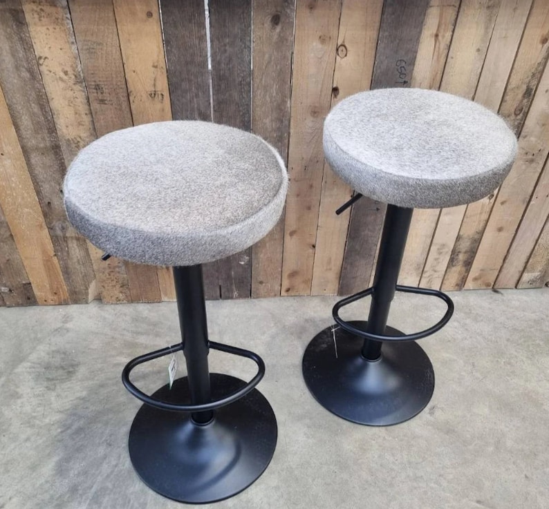 Op de afbeelding: Twee grijze en witte barstoelen met een zwart metalen onderstel en voetsteunen. De krukken zijn in hoogte verstelbaar.