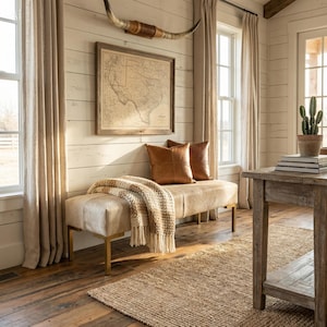 May include: A rectangular bench with a light-colored hide upholstery and gold-toned metal legs, topped with a textured cream and brown throw blanket and two brown leather pillows, positioned beneath a framed map and a longhorn steer skull.