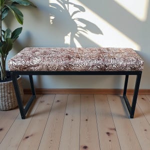 May include: A rectangular bench with a patterned cushion and a black metal frame. The cushion features a brown and white floral design. The bench is placed on a wooden floor, with a potted plant visible in the background.
