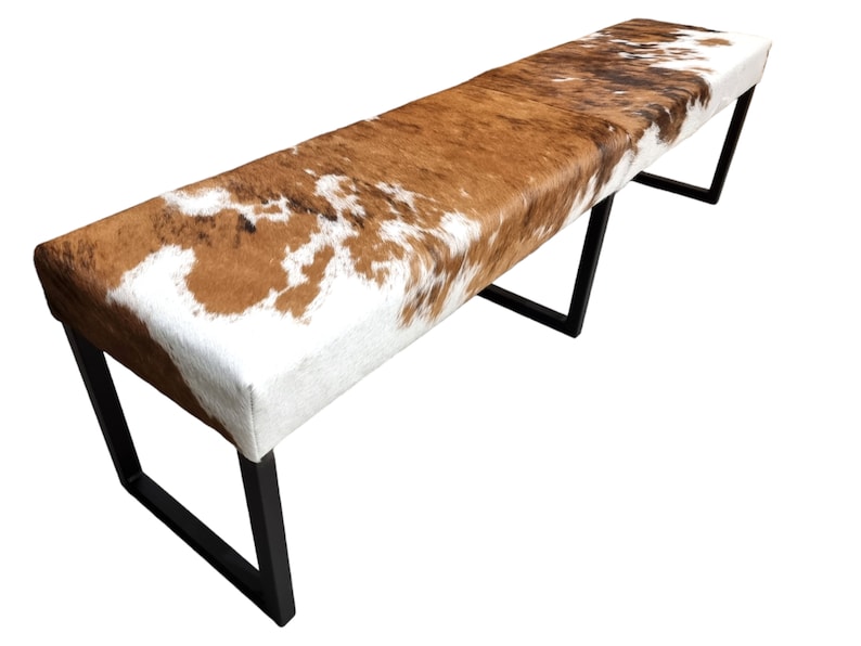 May include: A brown and white cowhide bench with a black metal frame. The bench has a rectangular shape and is supported by four legs.