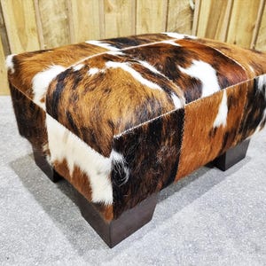 May include: A rectangular ottoman with a patchwork design made from cowhide. The cowhide features patches of brown, white, and black fur. The ottoman sits on four dark brown, square legs, offering a rustic aesthetic.