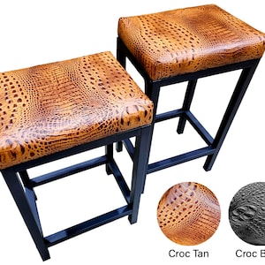 May include: Two bar stools with tan crocodile-patterned seats and black metal frames. The seats have a textured appearance, and the frames have a simple, modern design. The stools are shown against a white background.