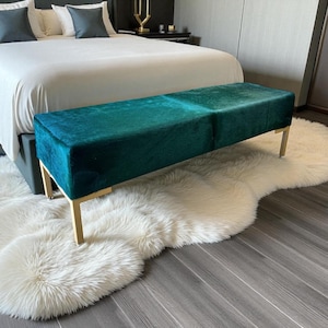 May include: A teal bench with gold legs sits at the foot of a bed. The bench is rectangular and upholstered in a plush, textured fabric. The bed is covered with a white comforter and pillows. A white, fluffy rug is beneath the bench.