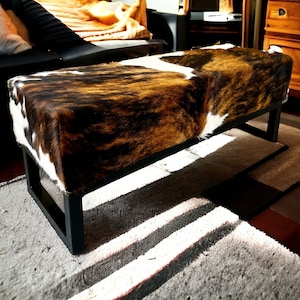 May include: A brown and white cowhide bench with a black metal frame. The bench is sitting on a rug with a white and gray pattern.