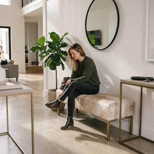 May include: A rectangular bench with a light-colored, mottled hide upholstery and a gold-toned metal frame. The bench is positioned in a bright room, featuring a smooth, light-colored surface and a sturdy, minimalist base structure.