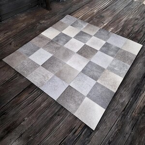 May include: A checkered rug made of cowhide in shades of grey and white. The rug is laid out on a wooden floor.