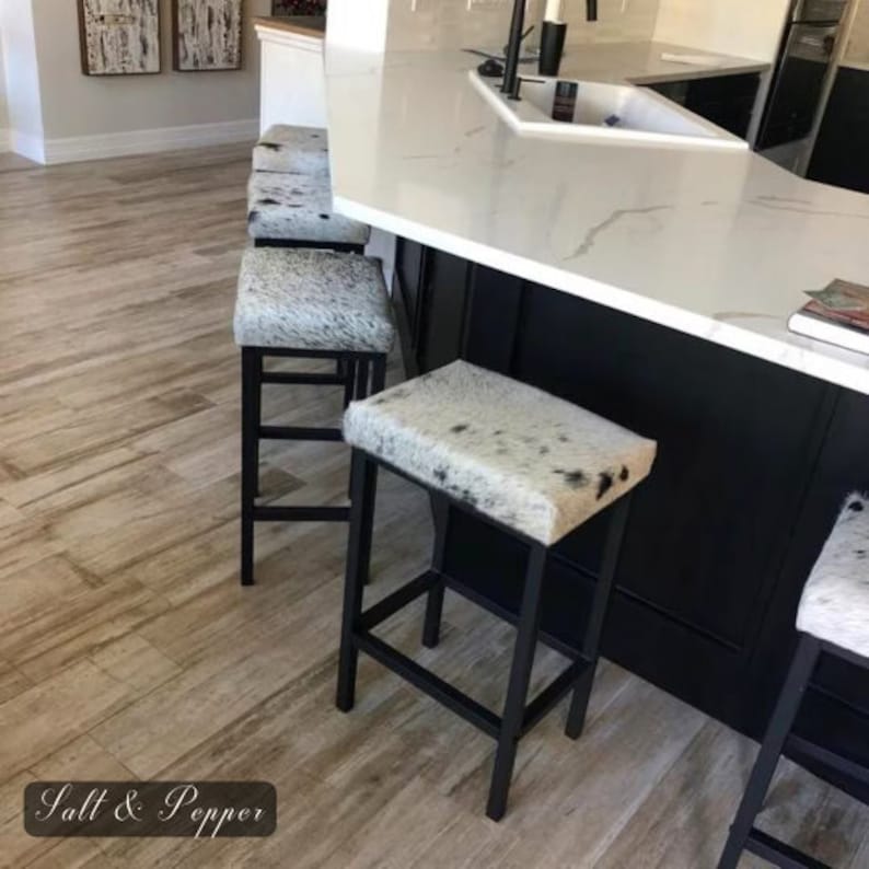 Peut inclure: Quatre tabourets de bar en m&eacute;tal noir avec des si&egrave;ges en peau de vache blanche et noire. Les tabourets sont dispos&eacute;s en rang&eacute;e devant un &icirc;lot de cuisine avec un plan de travail blanc.