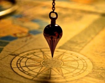 May include: A dark, metallic, teardrop-shaped pendulum hangs from a thin chain. The pointed tip of the pendulum is positioned directly above the center of a printed compass rose design on a textured, aged-looking map with warm yellow tones.