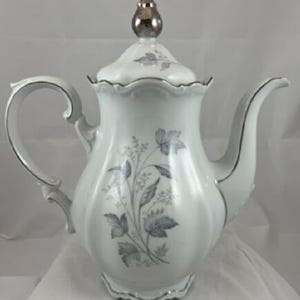 May include: White ceramic teapot with a silver-colored lid and trim. The teapot features a floral design in shades of gray and blue. The handle and spout are curved, and the teapot has a scalloped base.