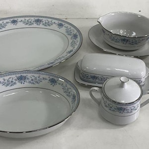May include: A collection of white porcelain dinnerware with a blue floral pattern and silver trim. The set includes an oval platter, a round serving bowl, a gravy boat with a saucer, a butter dish, a creamer, and a covered sugar bowl.