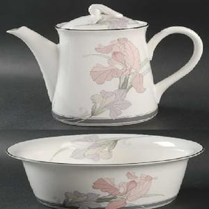 May include: A white porcelain teapot and matching bowl featuring a delicate floral pattern of soft pink and lavender iris flowers. Both pieces have a smooth, rounded silhouette with a thin silver trim along the base and rim of the bowl.