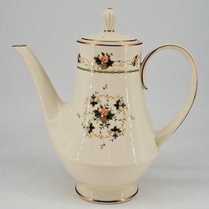 May include: Cream-colored ceramic coffee pot with a floral design. The pot has a long spout, a curved handle, and a lid with a small knob. Gold trim accents the edges and floral pattern. The floral pattern includes orange, blue, and yellow flowers.