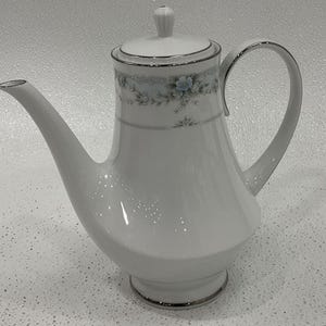 May include: White porcelain coffee pot with a floral design and silver trim. The pot has a curved spout, a large handle, and a lid with a small knob. The floral design features blue and green flowers.