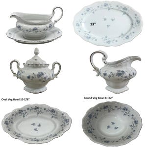 May include: A collection of white porcelain dinnerware featuring a blue floral pattern and silver trim. The set includes a gravy boat with a stand, a 13-inch oval platter, a lidded sugar bowl, a creamer, a 10 7/8-inch oval vegetable bowl, and an 8 1/2-inch round vegetable bowl.