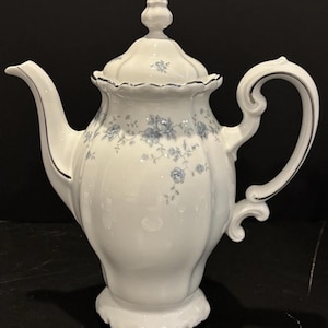 May include: White porcelain teapot with a floral blue pattern and silver trim. The teapot has a curved spout, a decorative handle, and a lid with a small finial. The design is classic and elegant.