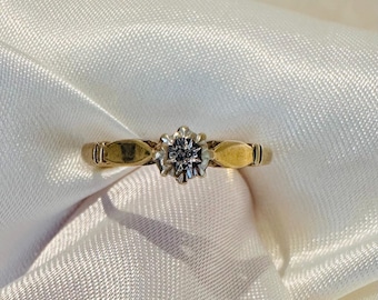 Könnte beinhalten: Ein goldener Ring mit einem blütenförmigen Mittelteil. Der Mittelteil hält einen kleinen, runden, klaren Edelstein. Das Band hat ein schlichtes Design mit zwei dekorativen Elementen auf beiden Seiten des Mittelstücks. Der Ring liegt auf weißem, strukturiertem Stoff.