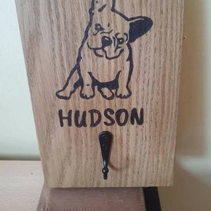 Puede incluir: Gancho de madera para la correa de un perro con un gancho de metal negro. El gancho está grabado con una silueta negra de un bulldog francés y el nombre "HUDSON".